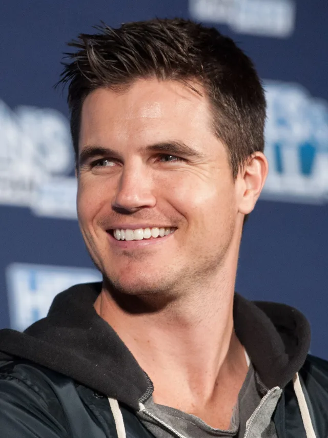 A smiling man with short dark spiky hair in a black hooded jacket, looking off to one side in front of a blue event backdrop.