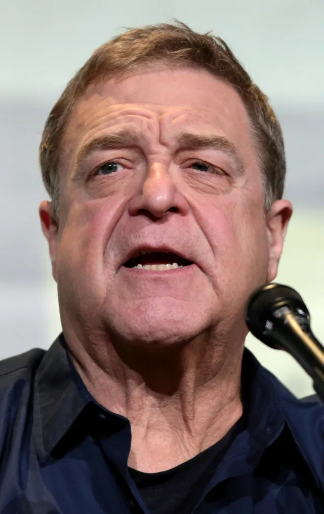 Close photograph of John Goodman speaking at a panel, in a dark blue shirt with a microphone at the edge of the frame.