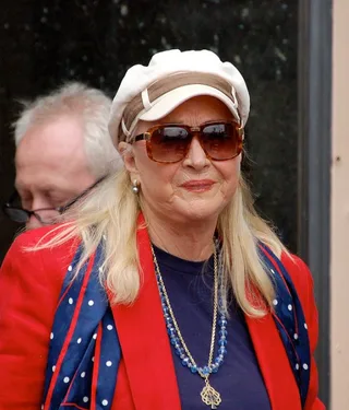 An older woman with long blonde hair, tinted sunglasses and a cream cap, wearing a red jacket with a blue polka-dot scarf and a beaded necklace.