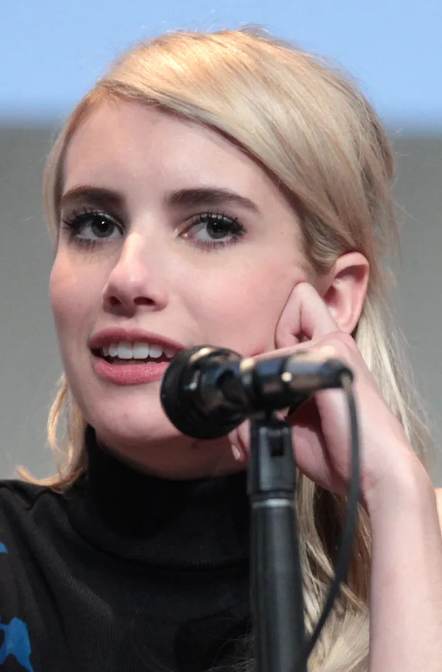 A woman with blonde hair swept to one side, resting her cheek on her hand and speaking, in a black polo neck with a microphone in front of her.