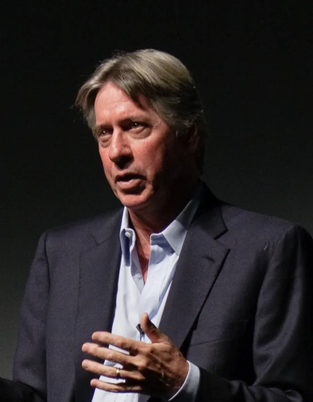 Alan Silvestri speaking on stage in a dark jacket and open-necked blue shirt, one hand raised as he talks.