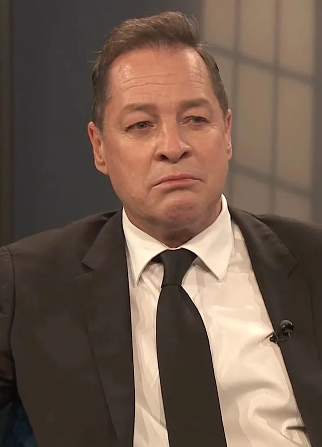 A man with short brown hair in a dark suit, white shirt and black tie, seated in a television studio with a clip-on microphone.