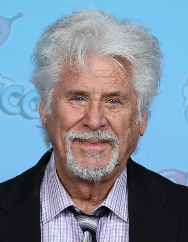 Older man with swept-back white hair and a grey goatee, wearing a dark jacket and checked shirt, smiling against a light blue backdrop.