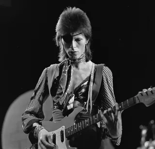 Black and white photograph of a musician with a feathered haircut and an eyepatch playing an electric guitar on a television stage.