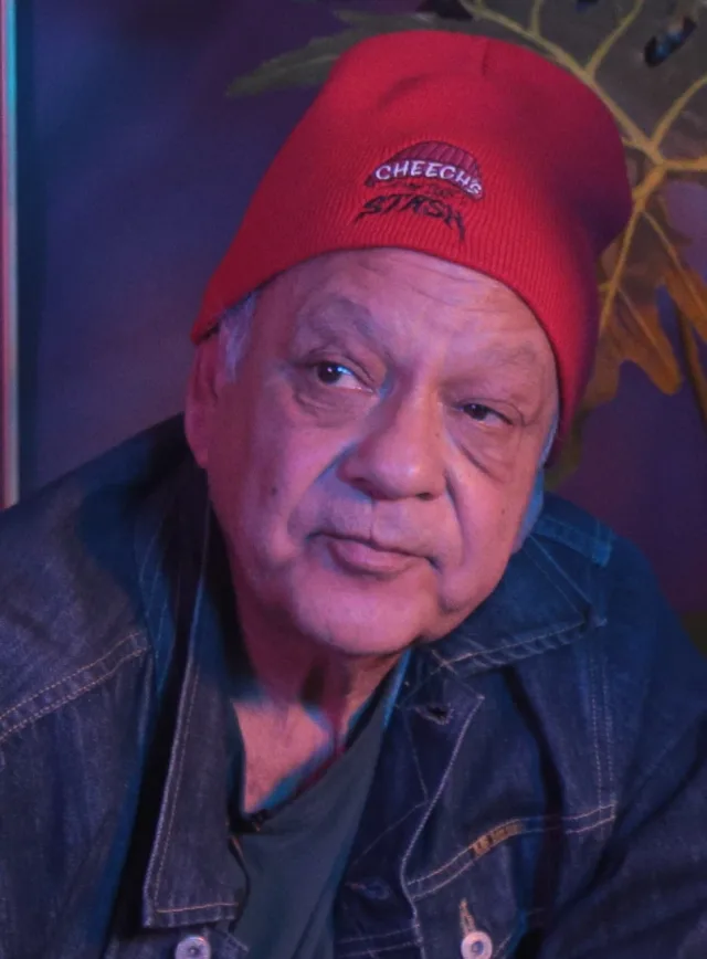 An older man in a red knitted beanie and a denim jacket, looking off to one side under purple lighting.