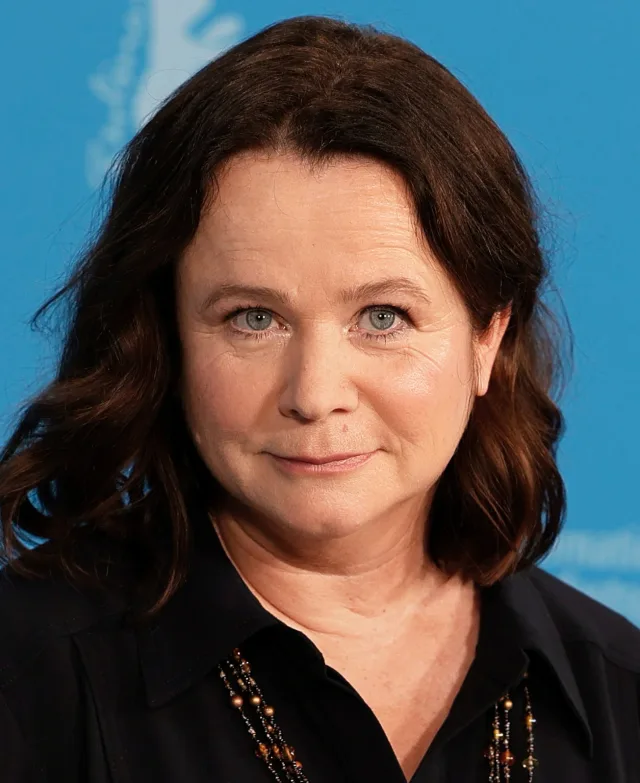 Close photograph of Emily Watson with shoulder-length dark brown hair, wearing a black collared top and a beaded necklace.