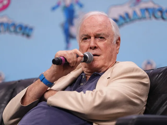 John Cleese speaking into a microphone on stage at a fan convention, seated in a cream jacket and navy shirt