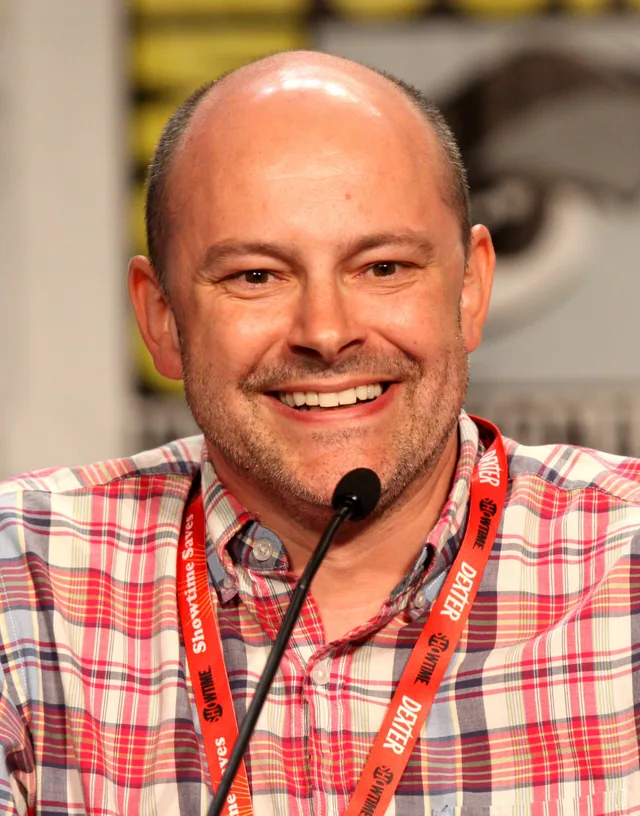 A smiling bald man with light stubble in a red and blue plaid shirt and an orange lanyard, seated behind a microphone at a convention panel.