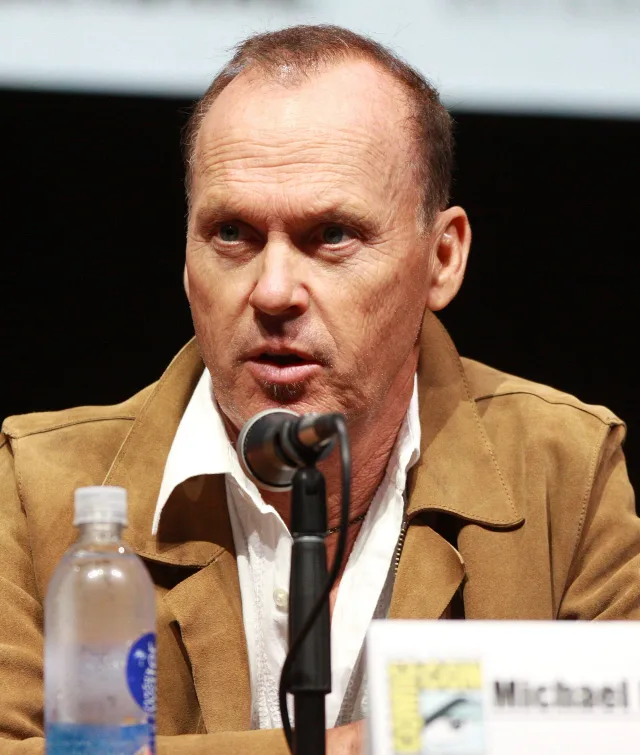 Close photograph of Michael Keaton speaking into a microphone at a convention panel, wearing a tan suede jacket over a white open-collared shirt.