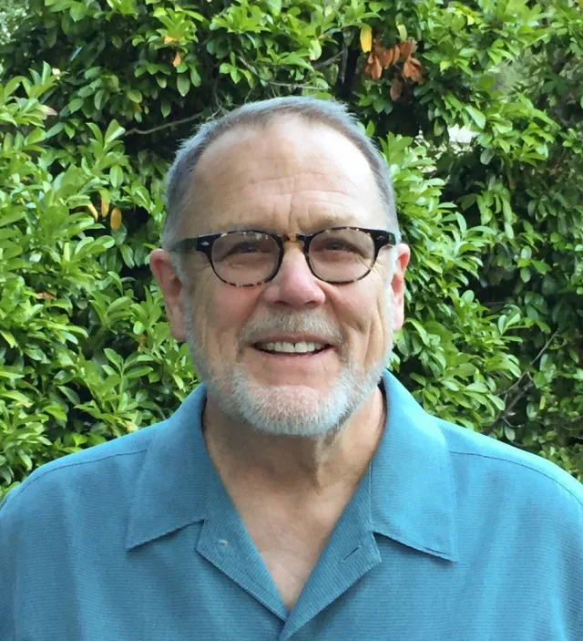 Smiling man with short grey hair, a grey beard and tortoiseshell glasses, in a teal shirt, standing in front of green foliage.