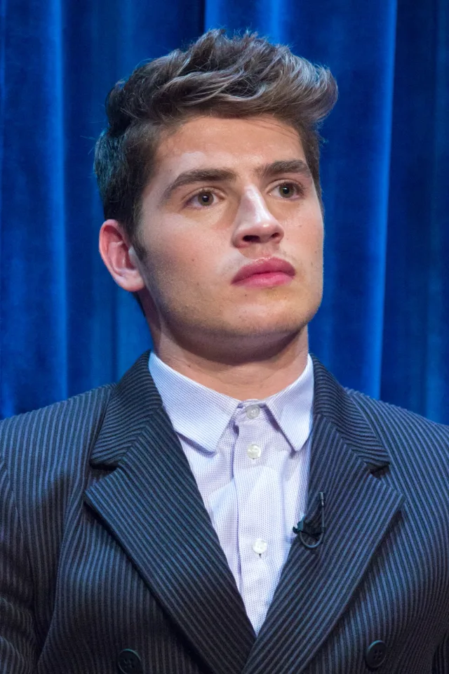 Gregg Sulkin with swept-up brown hair in a pinstriped jacket and lilac shirt, looking off to the side in front of a blue curtain.