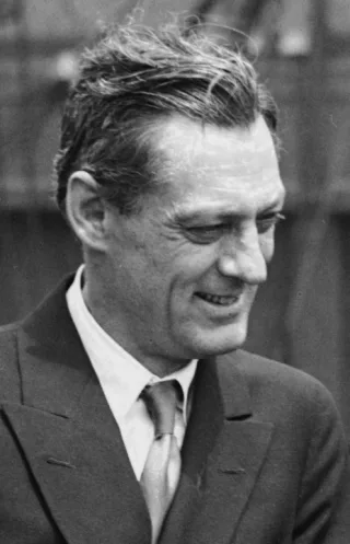 Black and white candid photograph of a man in a suit and tie, smiling and looking down to one side.