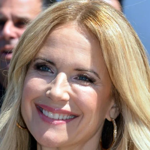 Close headshot of Kelly Preston smiling outdoors in bright sun, with long blonde hair and gold hoop earrings.