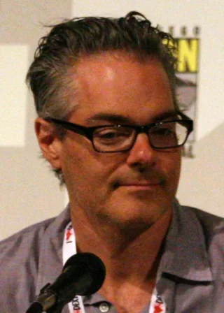 Close portrait of a man with swept-back grey hair and black-framed glasses, wearing a grey shirt and a lanyard, seated at a microphone at a convention panel.