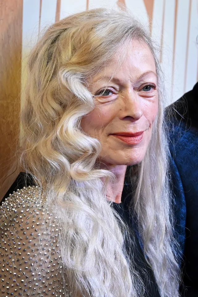An older woman with long, wavy blonde-grey hair and red lipstick, in a sparkly beaded top and dark blue wrap, looking off to the side.