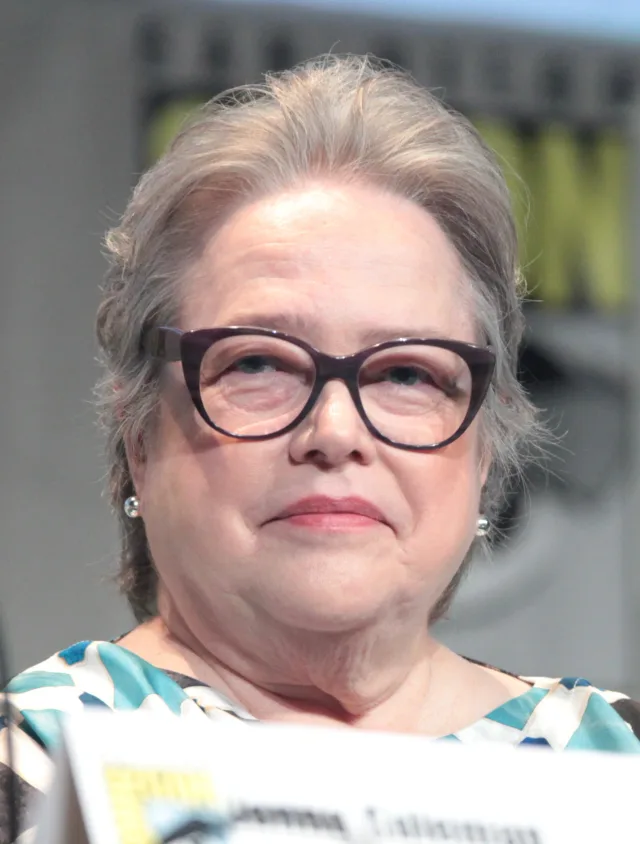 Close photograph of an older woman with grey swept-back hair, dark purple glasses and pearl earrings, smiling gently at a panel table.