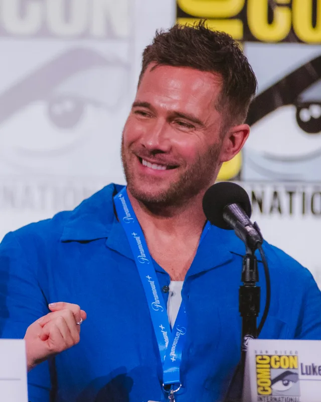 Smiling man with short brown hair and stubble, in a blue shirt, speaking at a convention panel table with a microphone.
