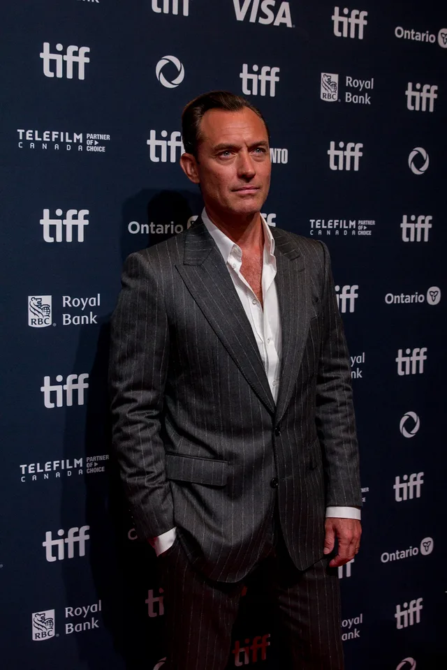A man in a grey pinstripe suit and open-necked white shirt, one hand in his pocket, standing in front of a dark blue sponsor wall at a film festival.