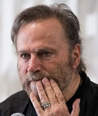 Portrait of an older man with swept-back grey-brown hair and a beard, hand raised to his chin, wearing a dark shirt, at a press event.