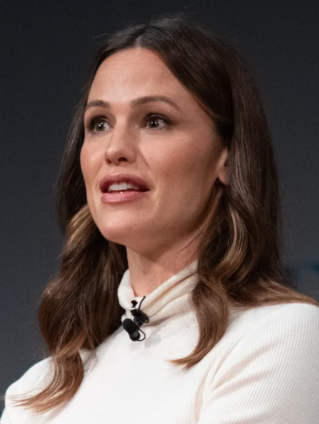 Close portrait of a woman with long brown hair, wearing a cream ribbed polo neck with a small clip microphone, speaking on a stage.