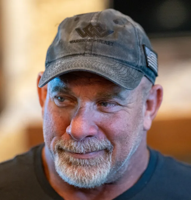Close photograph of a man with a grey beard in a dark cap and dark t-shirt, looking off to one side with a slight smile