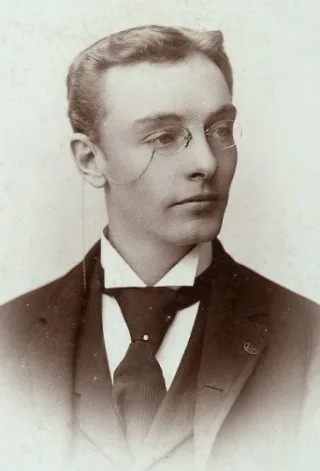 Sepia studio portrait of a young man in round wire glasses, a dark jacket and a striped tie, looking to one side.