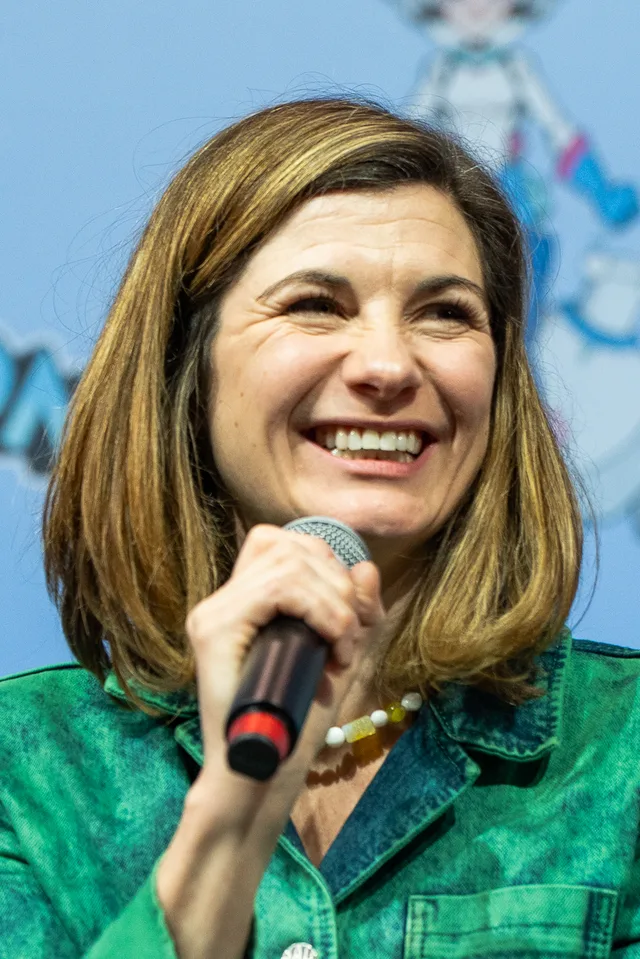 Laughing woman with shoulder-length brown hair holding a microphone, wearing a green denim jacket and a beaded necklace.