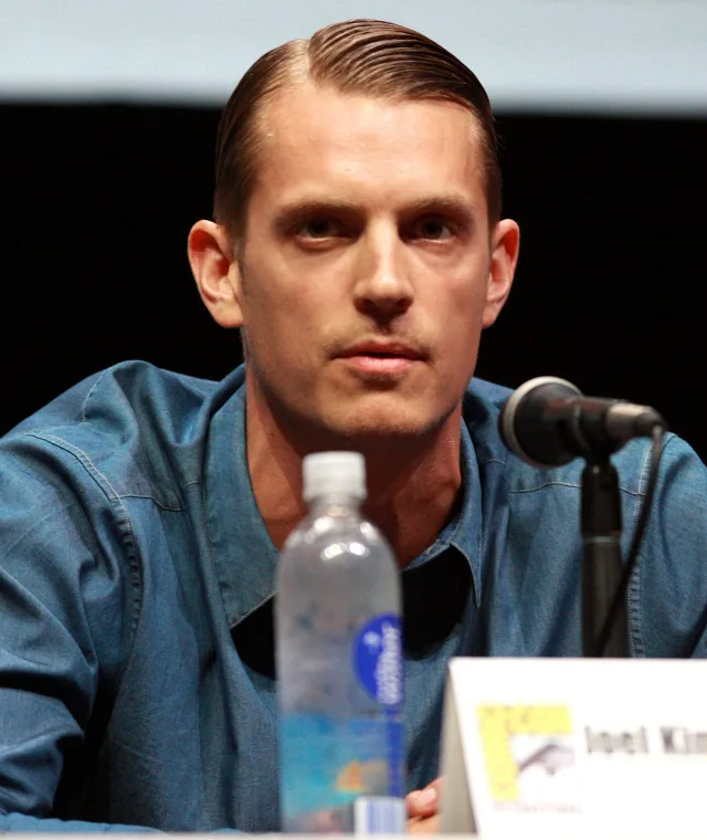 Joel Kinnaman in a blue denim shirt with slicked-back hair, seated at a panel table beside a microphone and a water bottle.
