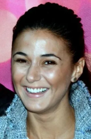 Close photograph of a smiling woman with dark hair pulled back, wearing a grey tweed jacket, against a pink background.