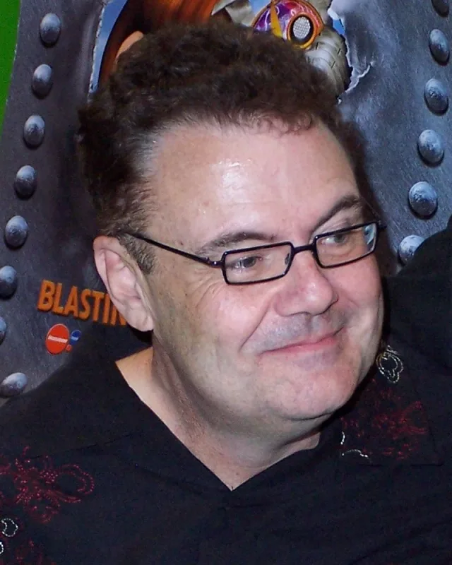Head and shoulders photograph of Glenn Shadix, a man with short dark curly hair and thin-framed glasses, smiling and looking to one side, in a dark top.