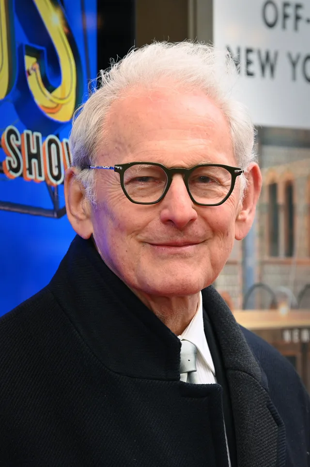 An older man with white hair and dark-framed glasses, smiling slightly, in a dark coat and pale tie on a city street.