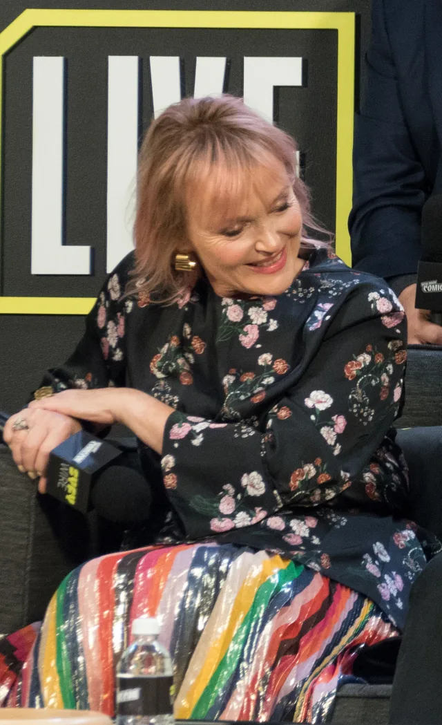 Smiling woman with a pink-tinted fringe and gold earring, looking down while seated on a panel sofa in a floral top and striped sequin skirt.