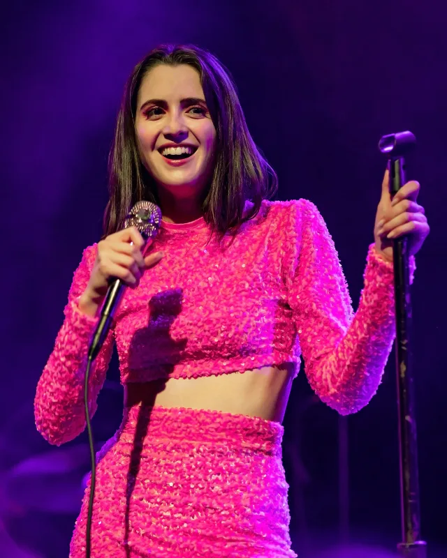 Laura Marano smiling on stage in a pink sequinned outfit, holding a microphone against a purple-lit background.