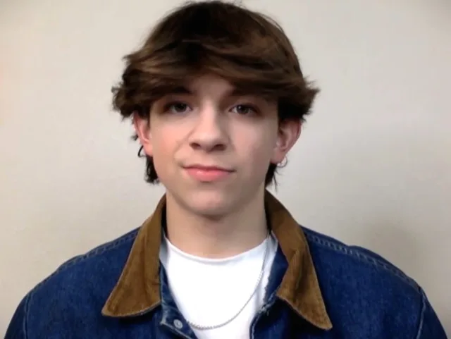 A teenage boy with brown tousled hair looking straight at the camera, in a denim jacket with a corduroy collar over a white t-shirt.