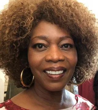 Close-up photograph of a smiling woman with voluminous curly golden-brown hair and large gold hoop earrings, in a red and white patterned top.