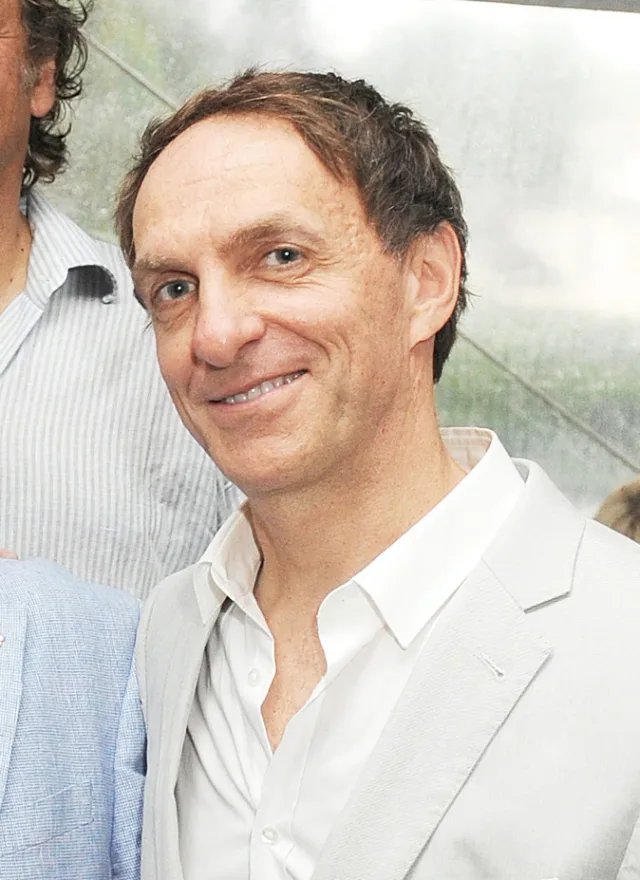 Smiling man with short brown hair in a light jacket and open-necked white shirt, other people partly visible at the edges.
