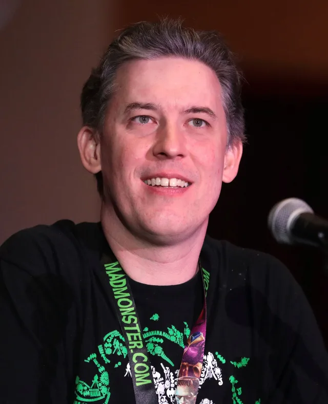 Smiling man with swept-back grey-brown hair in a black T-shirt and a green convention lanyard, speaking at a panel microphone.