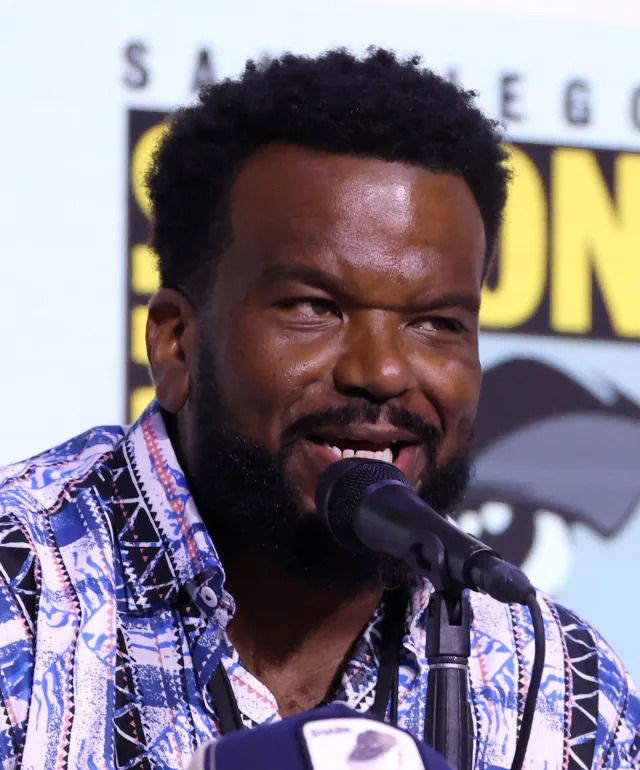 Craig Robinson laughing as he speaks into a microphone at a convention panel, wearing a blue and white patterned shirt.