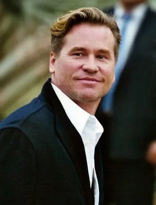 A man with swept-back blond hair and light stubble, smiling slightly, in a dark jacket over an open white shirt, photographed outdoors.