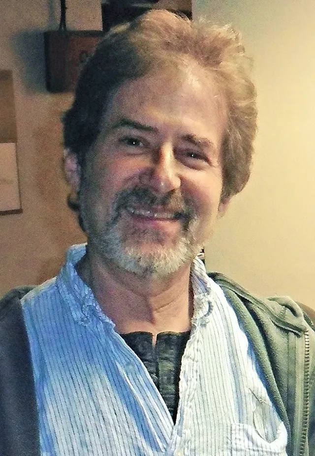 A smiling bearded man with tousled hair, wearing a light blue striped shirt under a green jacket, indoors.
