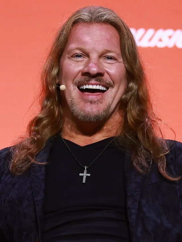 Chris Jericho laughing, with long wavy hair, a goatee and a small headset microphone, wearing a dark jacket and a cross necklace against an orange backdrop.