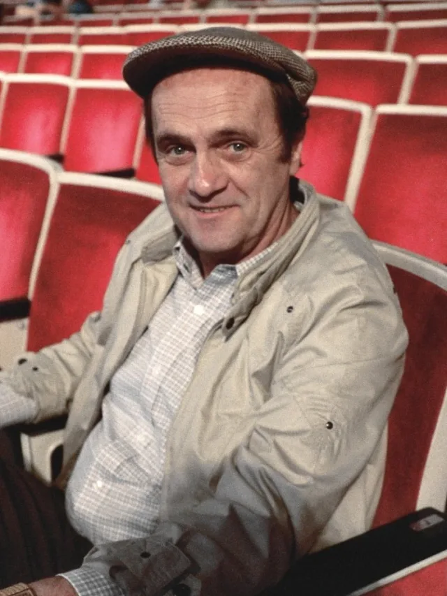 Bob Newhart smiling at the camera, seated in a theatre with rows of red seats behind him, wearing a flat cap and a light jacket.