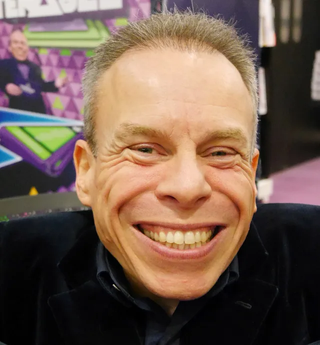Warwick Davis grinning broadly at the camera, wearing a dark velvet jacket and dark shirt at an indoor event