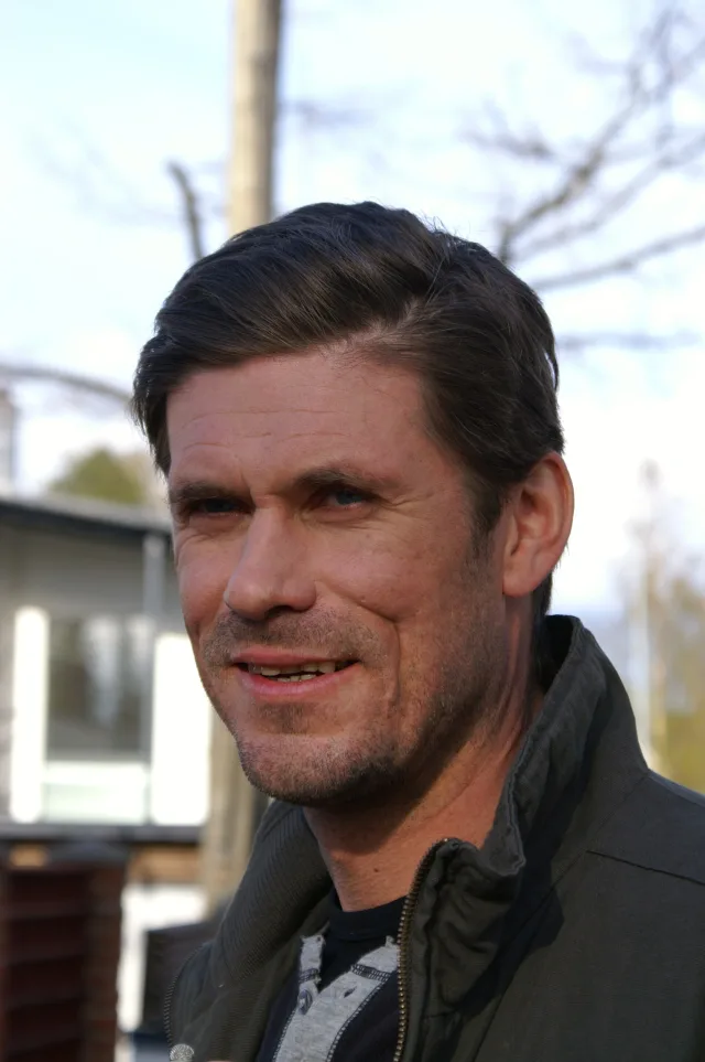 Smiling man with swept-back dark hair and stubble, outdoors in a dark zip jacket