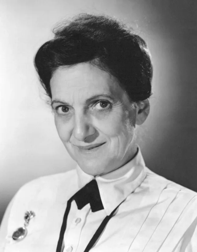 Black and white studio portrait of an older woman with dark swept-up hair, a gentle half smile and a white pleated blouse with a dark bow collar and a brooch watch.