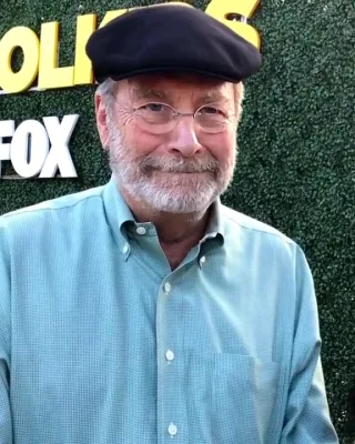 Martin Mull, a bearded older man in round glasses, a dark flat cap and a green checked shirt, smiling at the camera.
