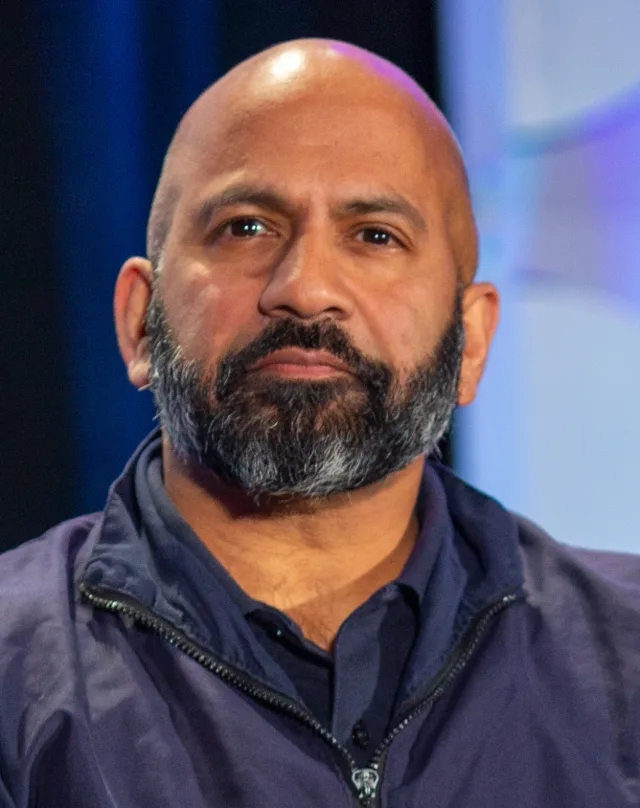 Close photograph of a bald man with a grey-flecked beard in a navy zip-neck jacket, seated against a blurred blue background at a panel event.