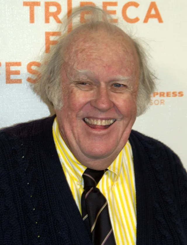 A smiling elderly man with tufts of grey hair, in a navy cardigan and a yellow striped shirt, in front of a festival backdrop.