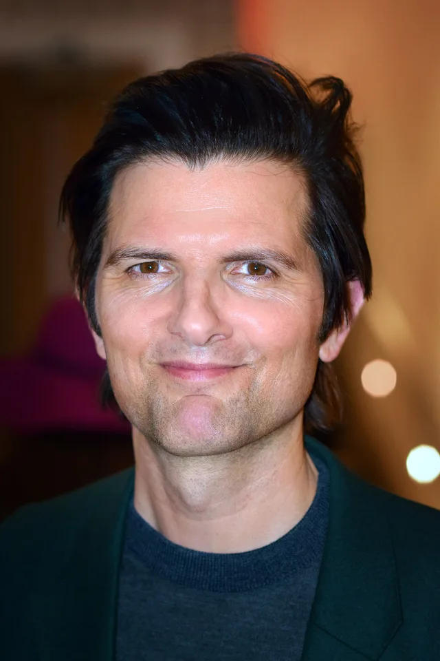 Close portrait of a smiling man with dark swept-back hair and stubble, wearing a dark green jacket over a grey sweater, with soft blurred lights behind him.
