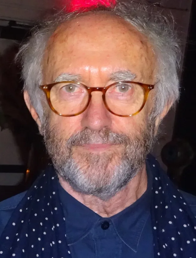 Close portrait of an older man with untidy grey hair, a grey beard and round tortoiseshell glasses, wearing a navy polka-dot scarf.